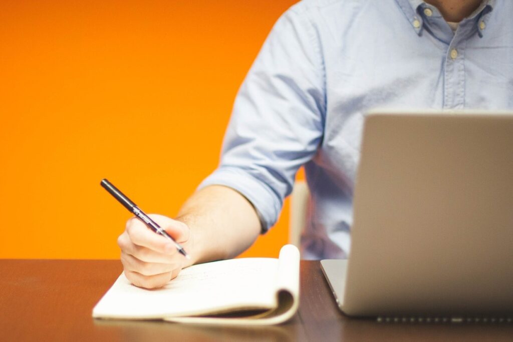 man at a laptop, writing in a notebook