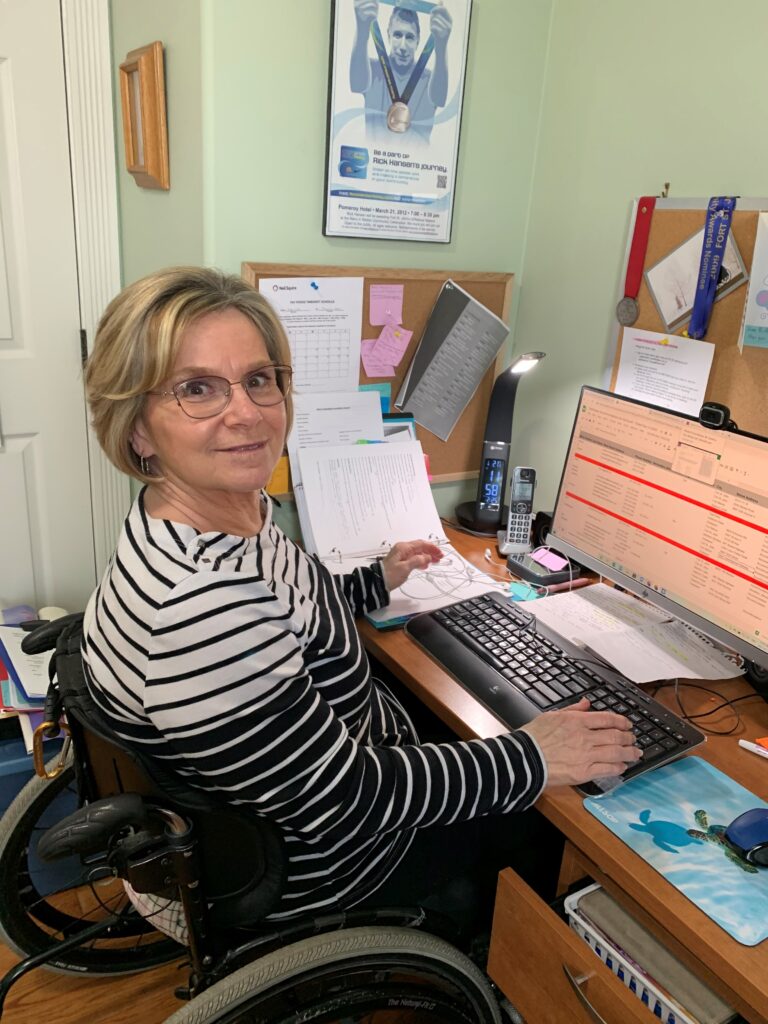 Lori working at her desk, seated in her wheelchair, looking at the camera and smiling