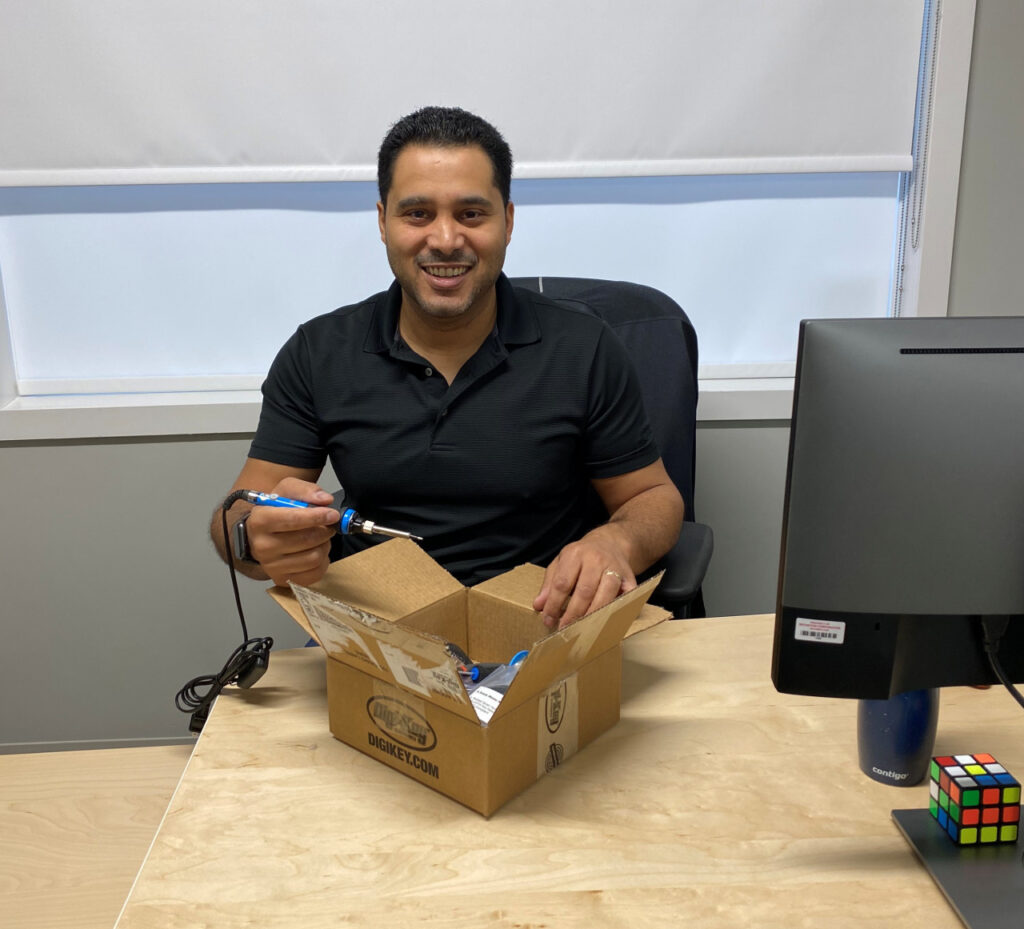 Dan Belhassen, president of Neovation, smiles and holds a soldering iron, as he holds open a box of parts to build a Round Flexure Switch