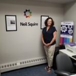 Staff member Tracey posing and smilin next to a Neil Squire banner, with a Neil Squire logo on the wall behind