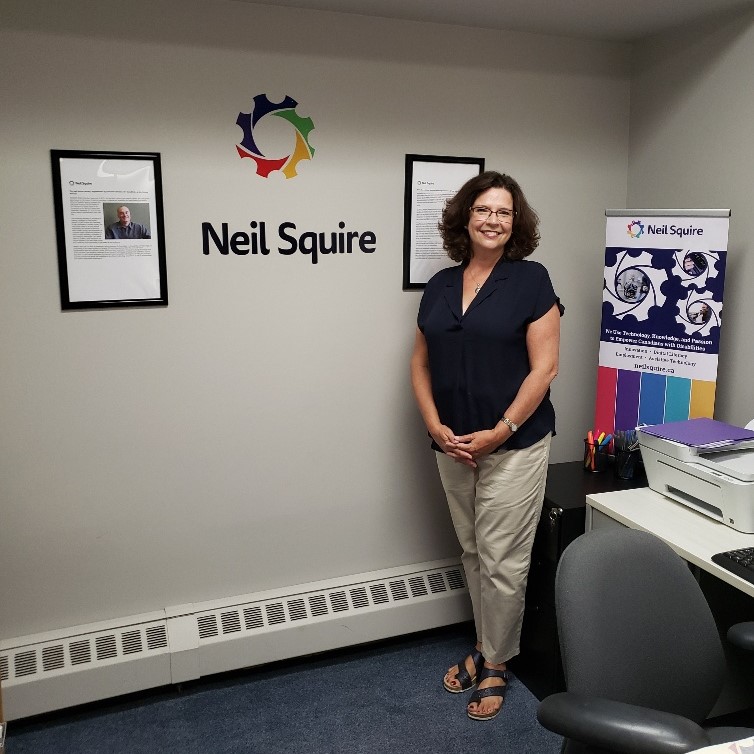 Staff member Tracey posing and smilin next to a Neil Squire banner, with a Neil Squire logo on the wall behind