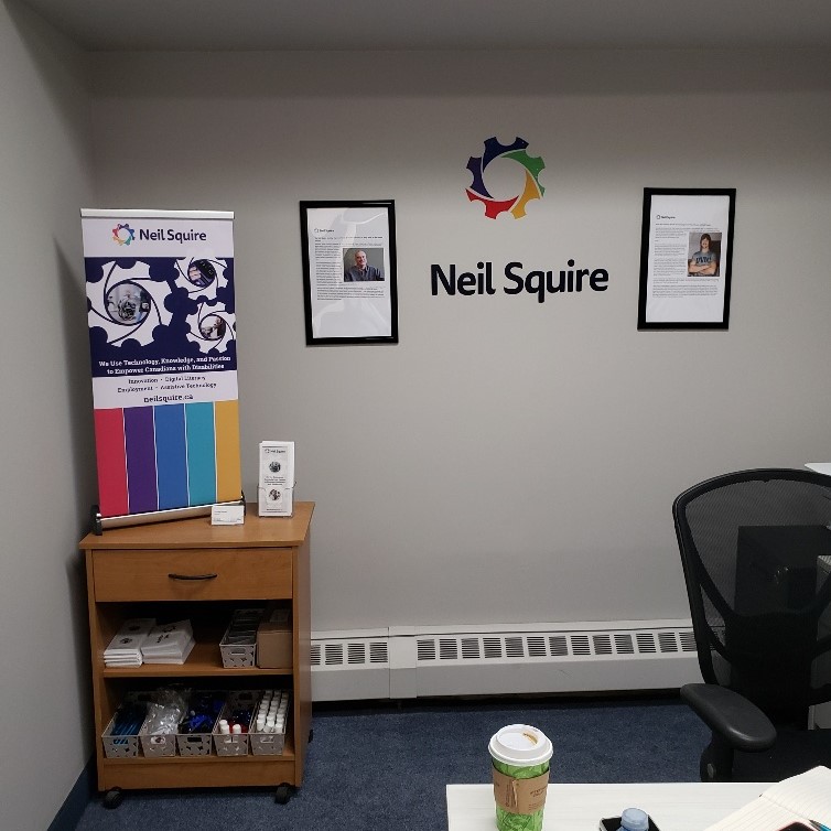 A small table with a Neil Squire banner on it. On the wall is a Neil Squire logo and two framed articles: one of Neil Squire, the other of Gary Birch
