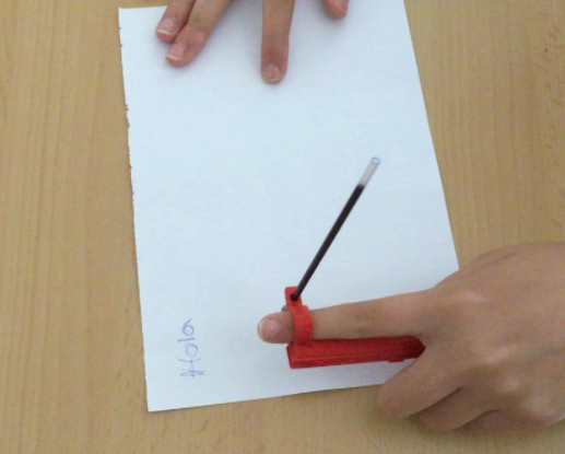 A person uses the Arm-mounted Pen Holder to write "Hola" on a piece of paper twice