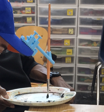 a person uses an adapted head pointer attached to their baseball cap to dip a paintbrush in paint