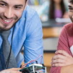 a teacher and two students chatting and laughing as the teacher demonstrates an electronic device