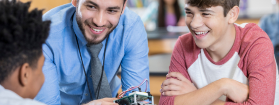 a teacher and two students chatting and laughing as the teacher demonstrates an electronic device