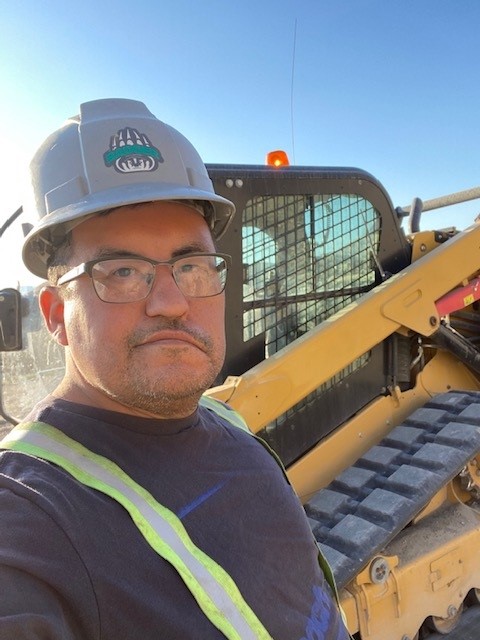 Bluejay wearing a hard hat and work clothes in front of heavy machinery