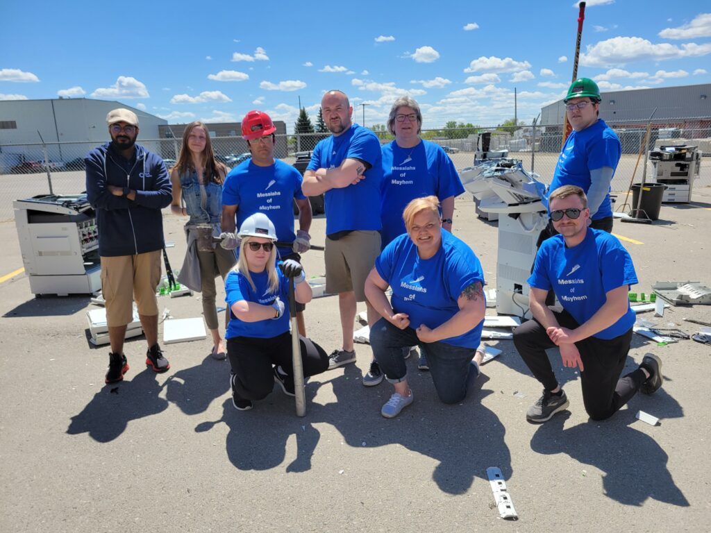 Neil Squire Prairie Region employees standing posing together wearing "Messiahs of Mayhem" shirts in front of smashed up photo copiers