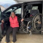Joan sitting in her adapted van with the door open, with her wheelchair being loaded into her van