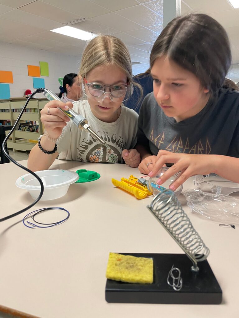 two girls working together on a device, with one holding a soldering iron