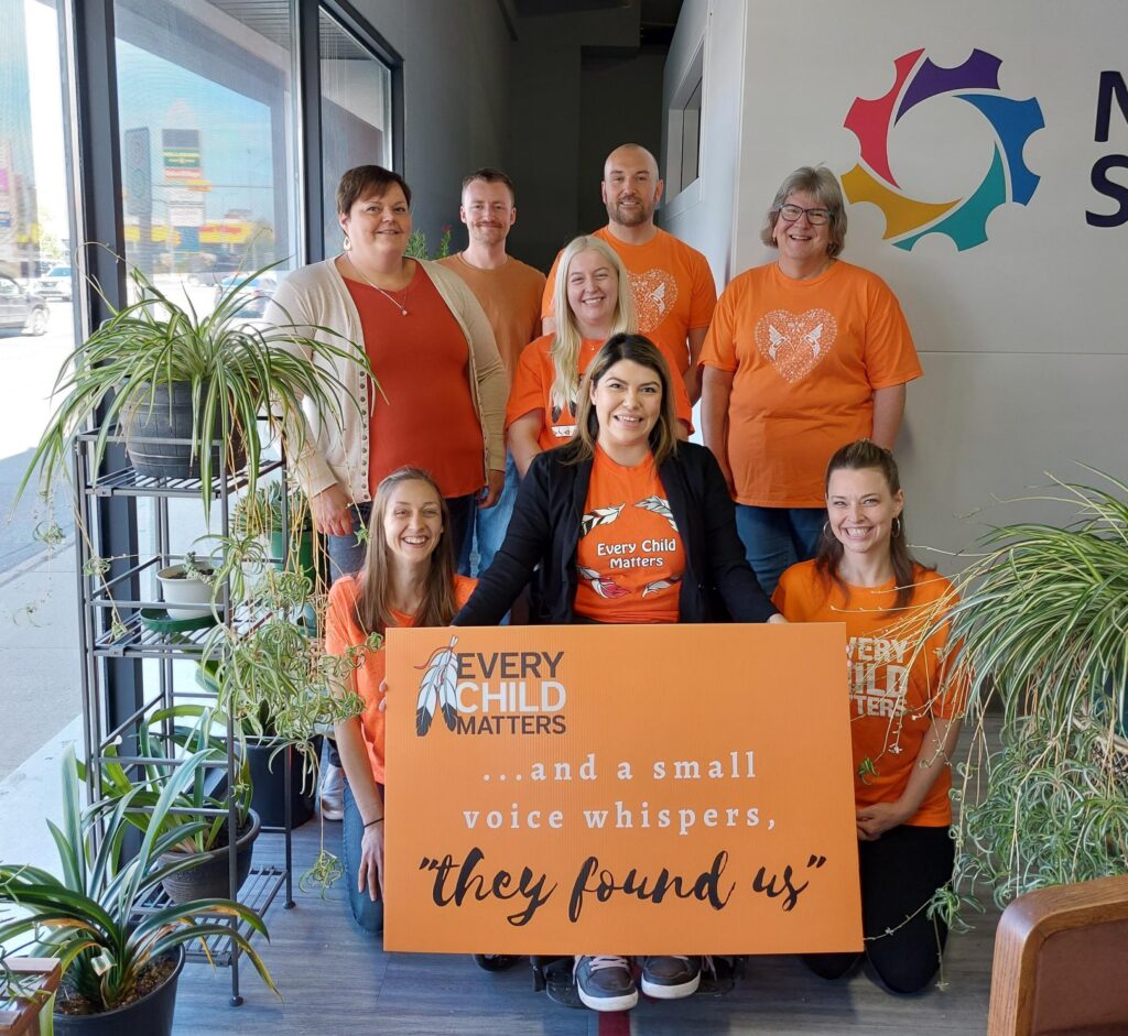 Neil Squire Regina staff wearing orange shirts and a sign that says, "Every Child Matters. ...A small voice whispers, "They found us"