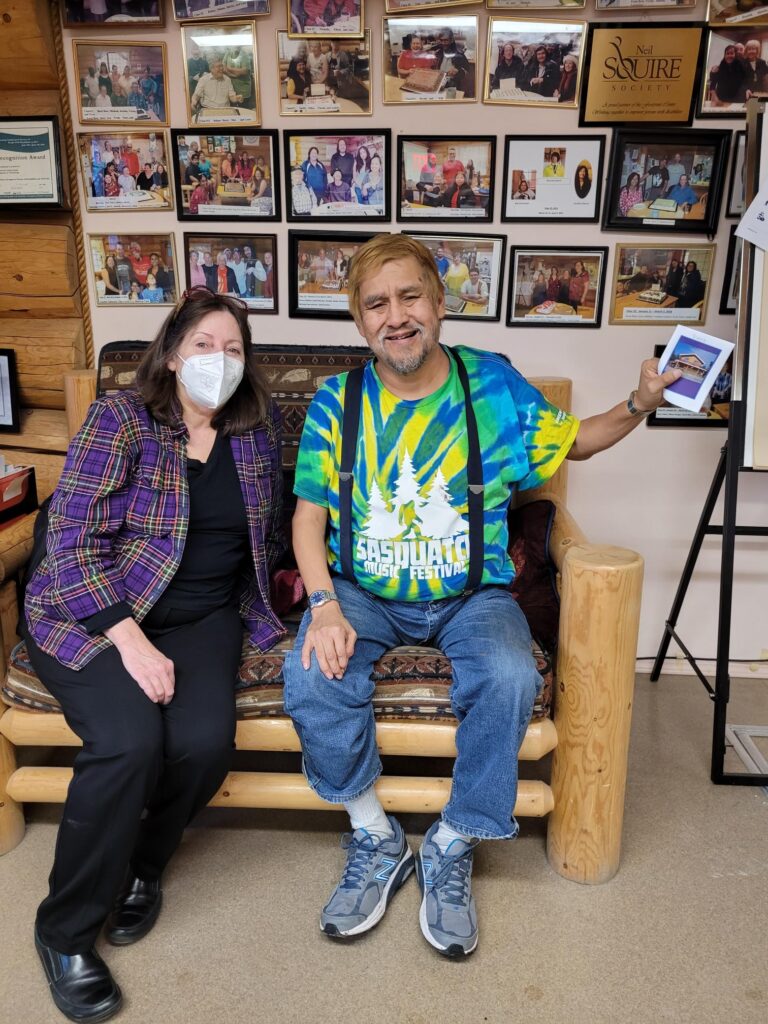 Footprints participant Vince sitting on a couch in front of wall of photos, next to Mindy, who helps run the program