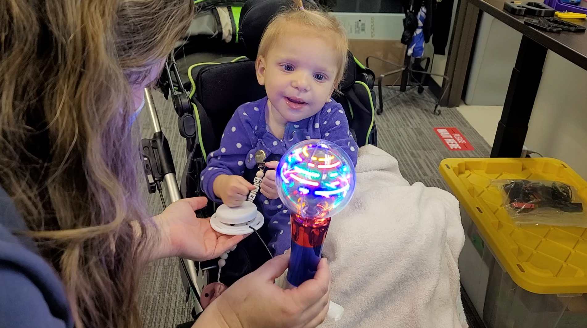 A toddler girl in a stroller playing with a spinning light wand with an assistive switch. The toy and switch is held by her mom on the left