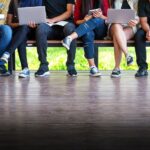 various people sitting on a bench using laptops and tablets
