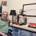 Alfred in a wheelchair sitting in front of his 3D printer and LipSync casings he has printed. A sign for his business, &quot;Accessible Places,&quot; hangs in the background.