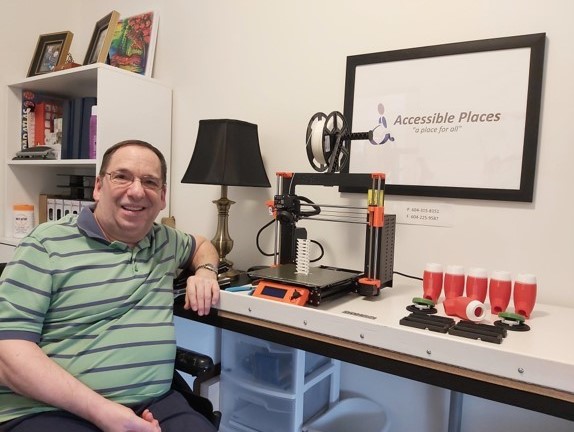 Alfred in a wheelchair sitting in front of his 3D printer and LipSync casings he has printed. A sign for his business, &quot;Accessible Places,&quot; hangs in the background.