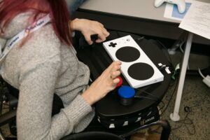A person in a wheelchair plays a video game using an Xbox Adaptive Controller and Assistive Switches on her wheelchair tray.
