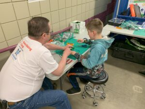 A man wearing a Makers Making Change shirt helps a kid solder at a build event.