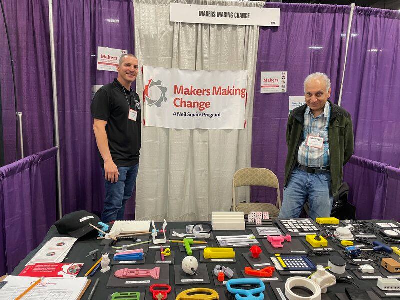 Farrukh and Christopher, MMC New York City Chapter Leaders, standing in front of a table with various 3D printed assistive tech