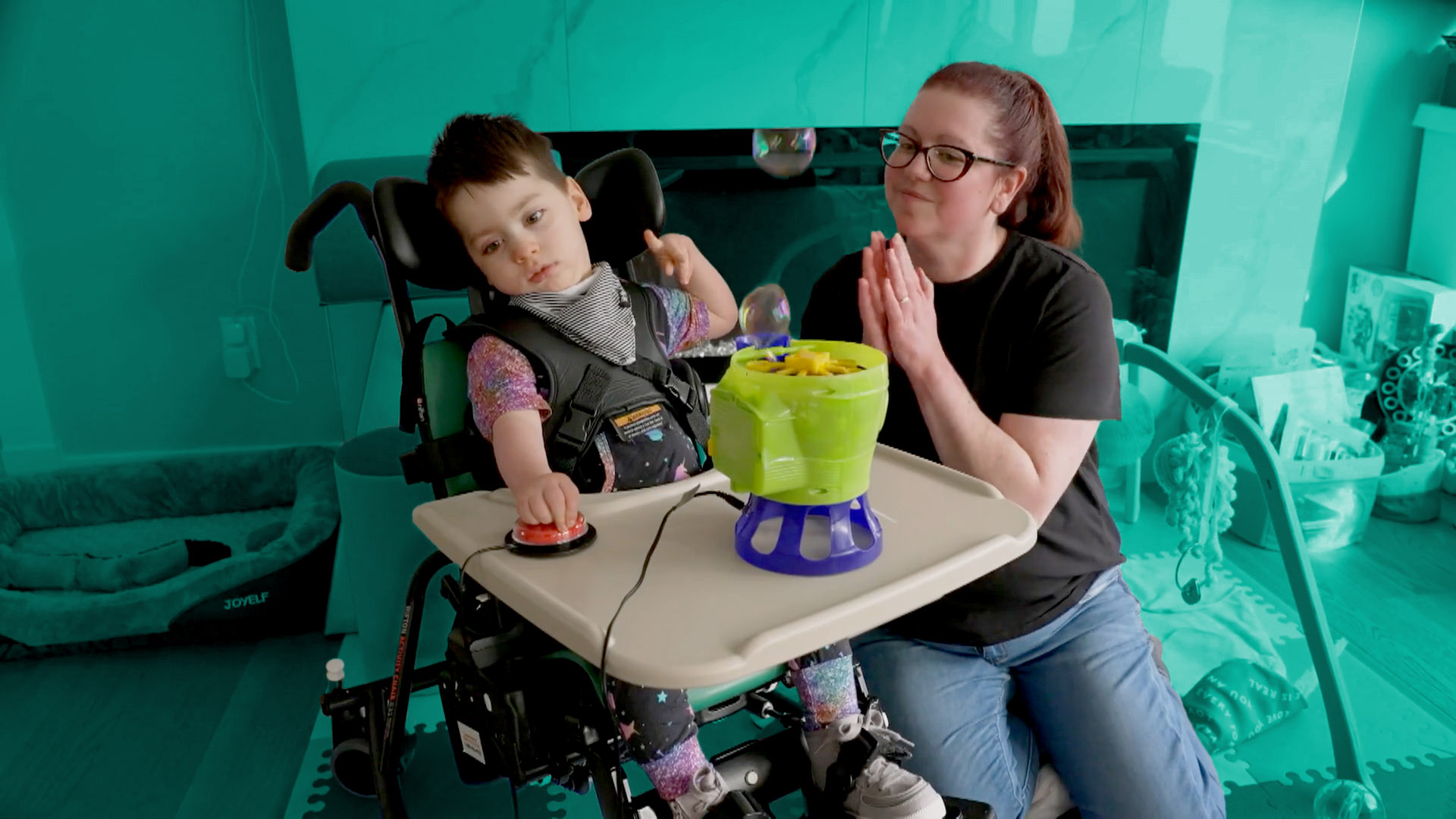 A woman watches as a child in a wheelchair uses a switch-adapted bubble blower toy.