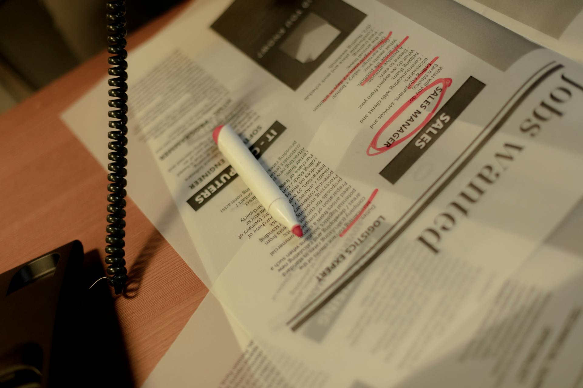 A job seeker uses a phone and circles job ads in the newspaper using a red marker.