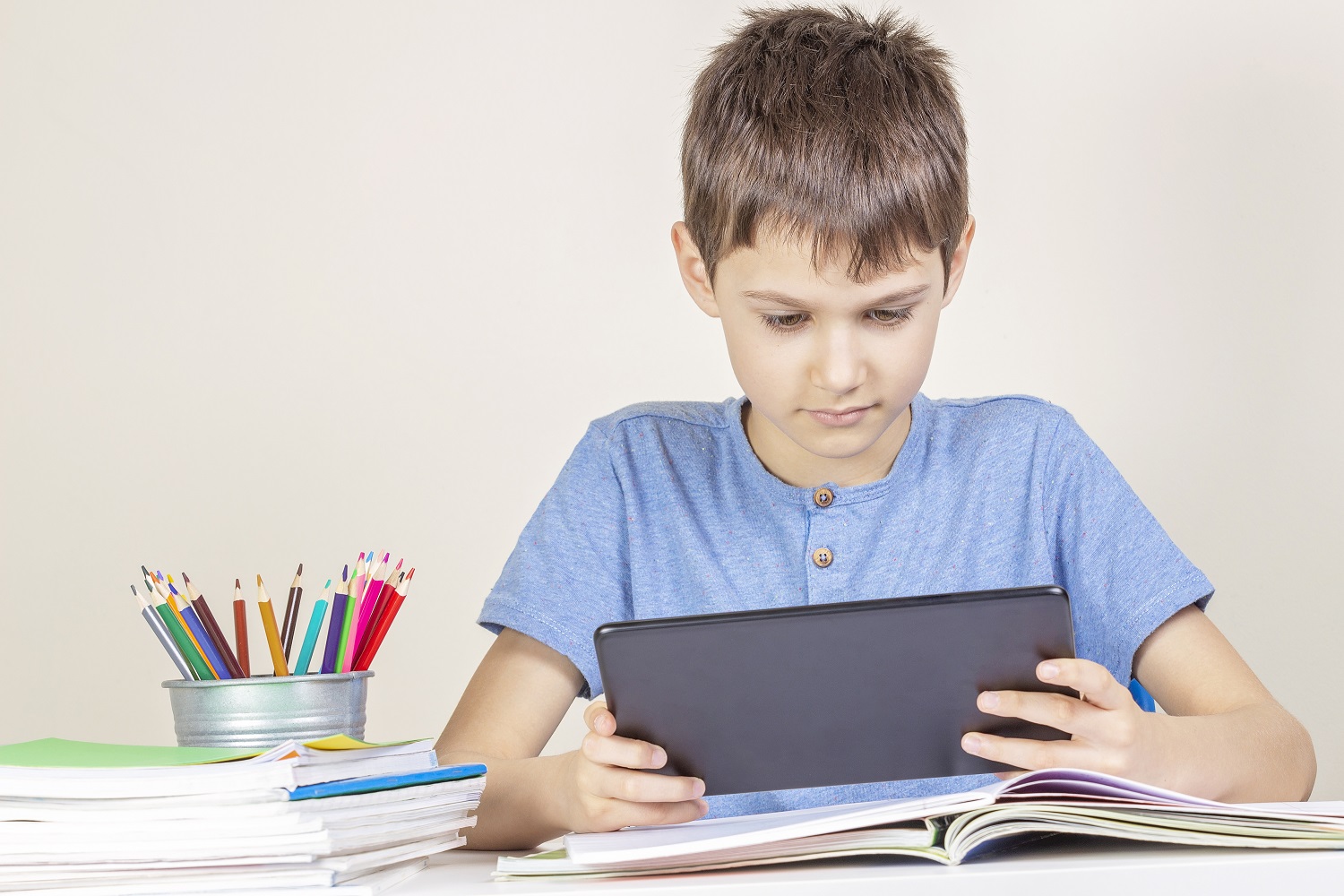 A kid sits down at a table with books and pencils on it, and uses a tablet.