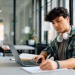 A student takes notes while going on a laptop.