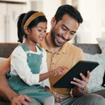 A father holds a tablet as his daughter points at the screen.
