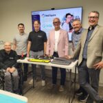 Gary, Eric, Justin, MP Ernie Klassen, Nate, and Chad smile together in a group photo behind a table with assistive technology on it.