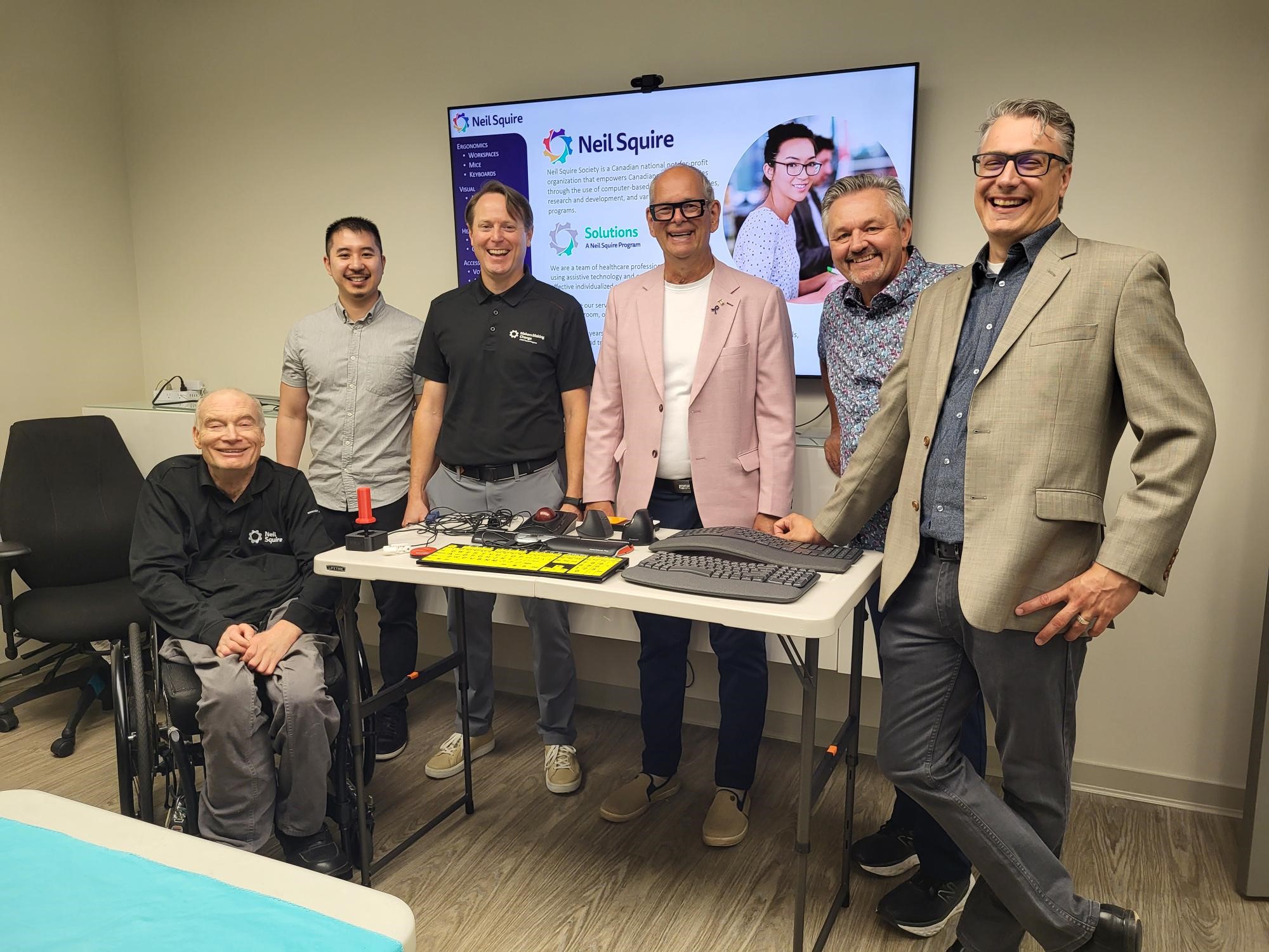 Gary, Eric, Justin, MP Ernie Klassen, Nate, and Chad smile together in a group photo behind a table with assistive technology on it.