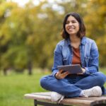 A woman sits cross-legged on a bench outside holding a tablet.
