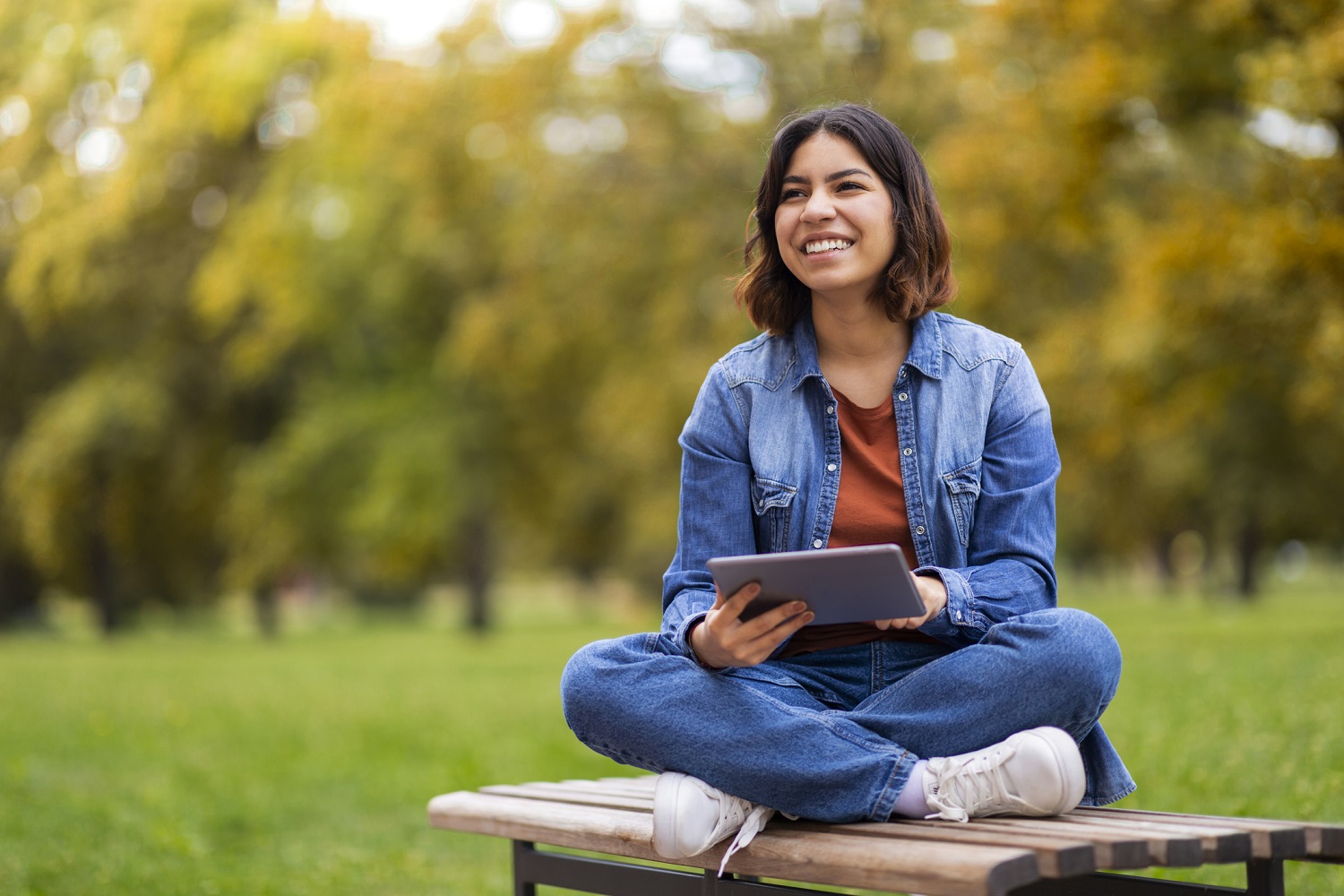 A woman sits cross-legged on a bench outside holding a tablet.