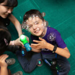 A young boy plays with bubbles from a switch adapted bubble blower, which is held by a pair of clinicians.
