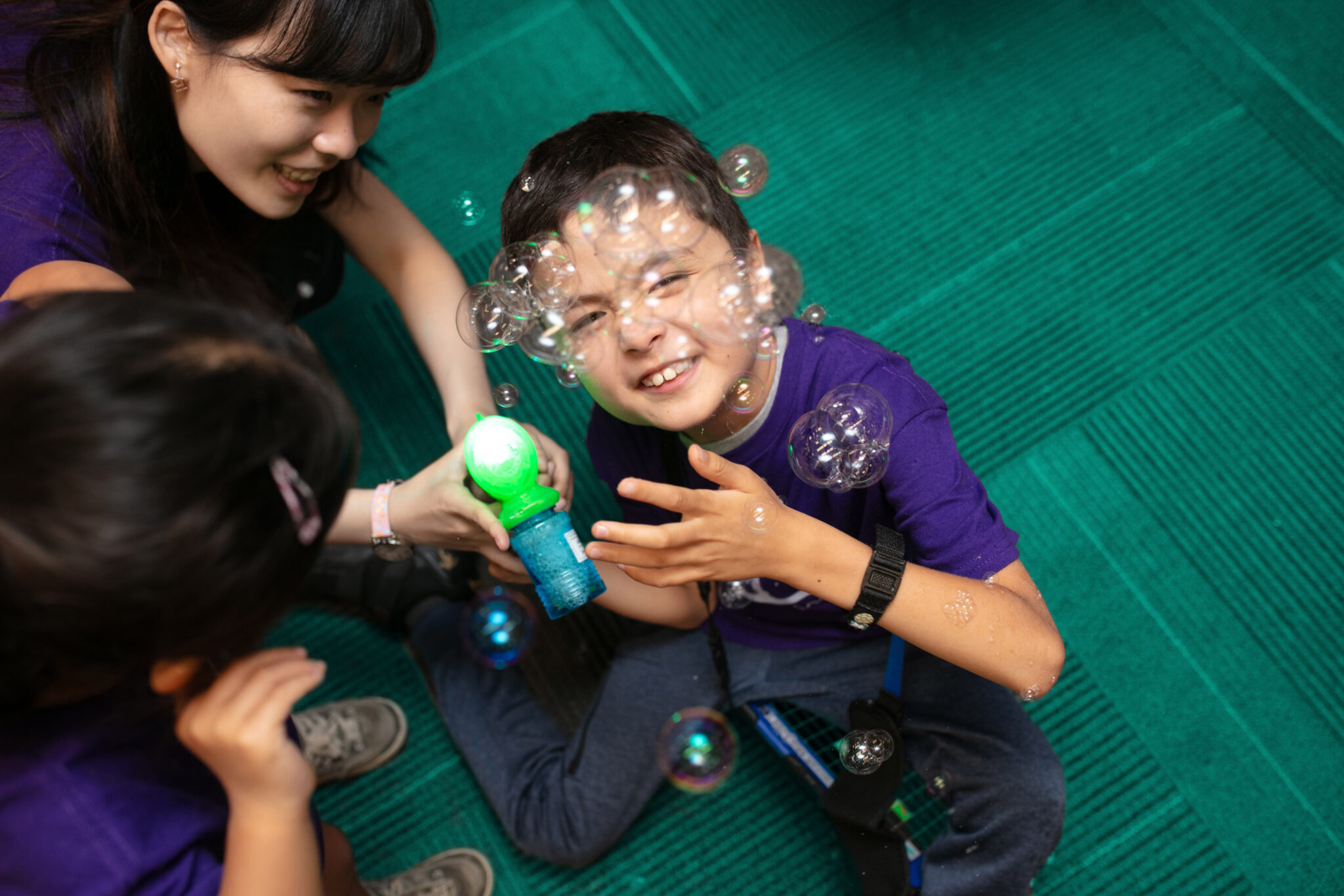 A young boy plays with bubbles from a switch adapted bubble blower, which is held by a pair of clinicians.