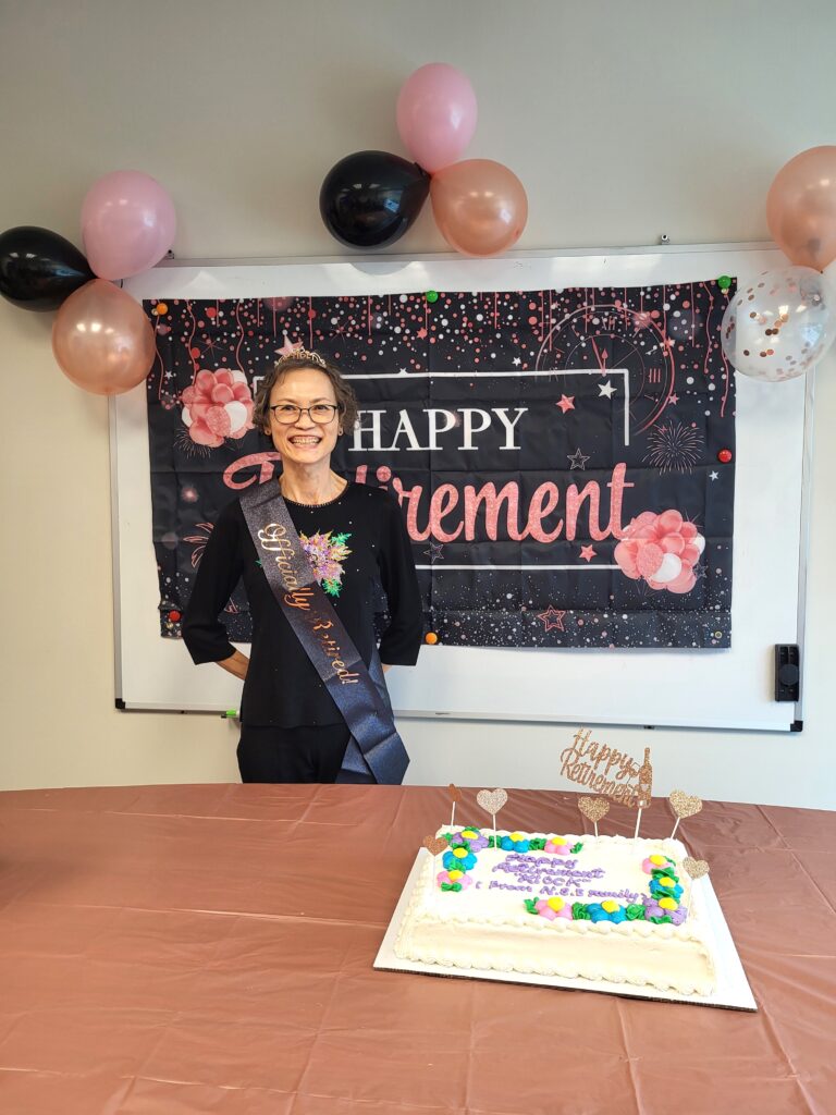 Kiock in front of a happy retirement banner, balloons, with a cake in front of her.