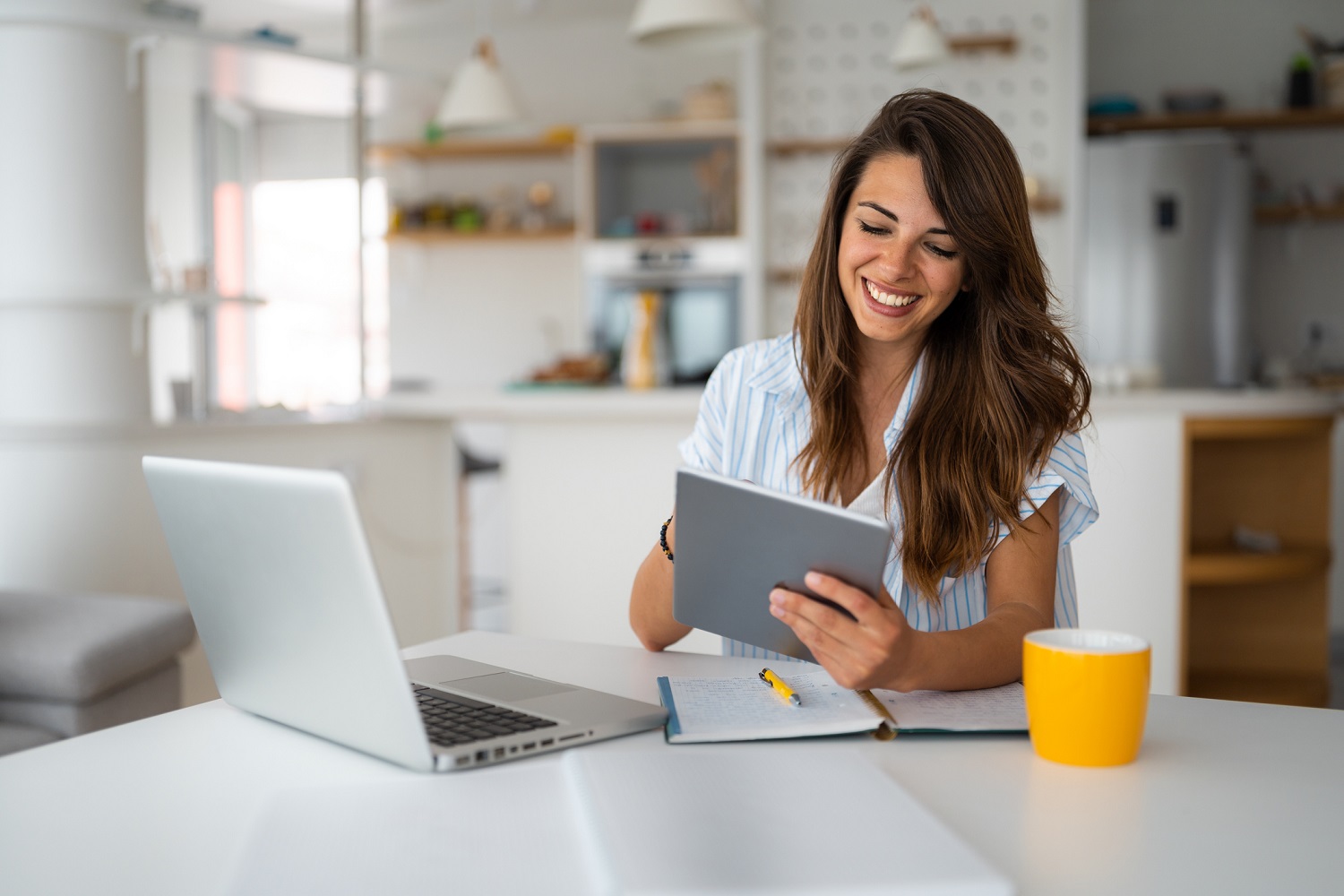 A woman sitting near a notepad pen, uses a tablet while also having a laptop open.