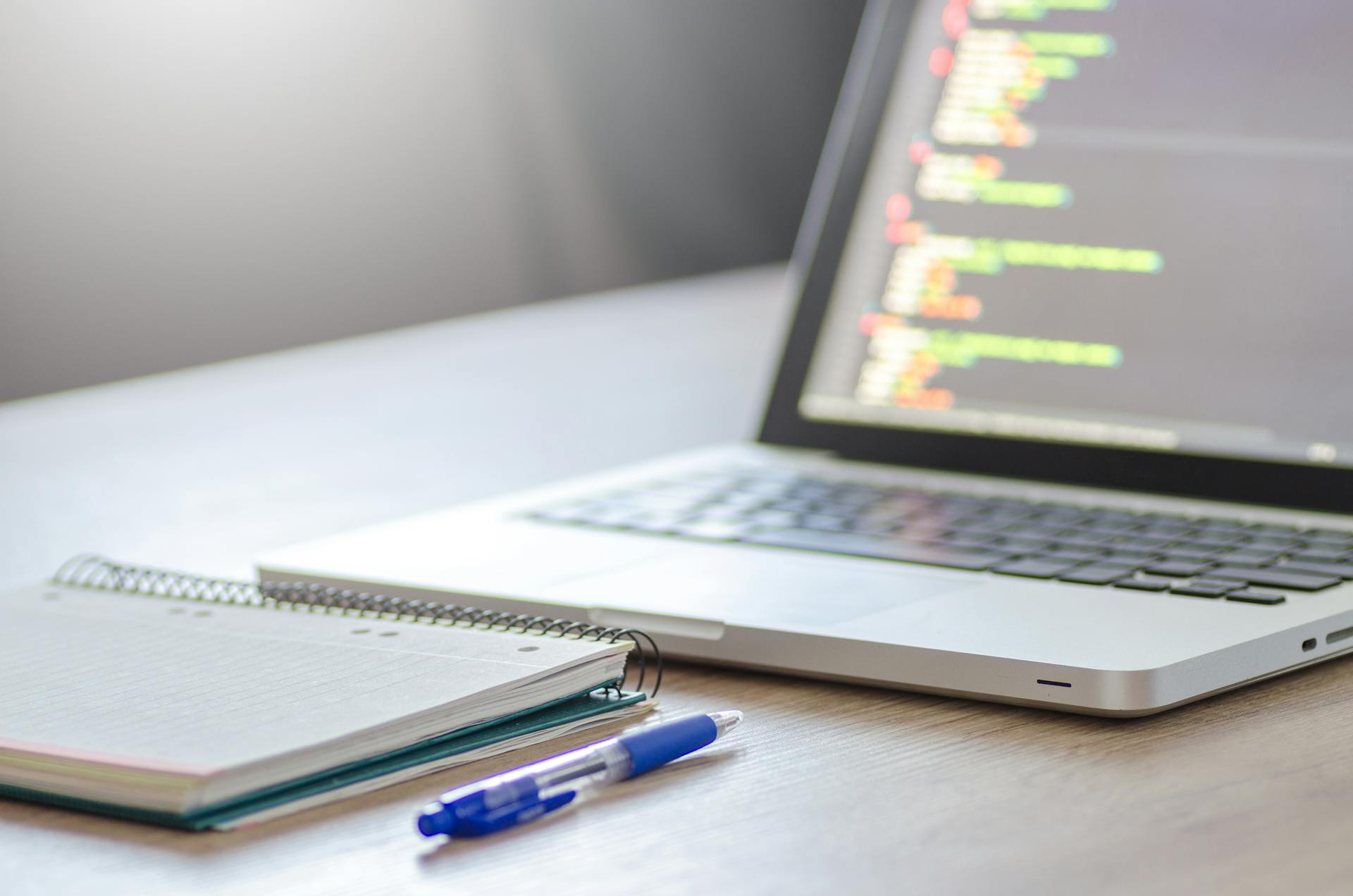 A laptop being used for coding, with a notebook and pen in front of it.