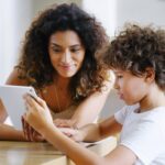 An adult woman watches as a young boy uses a tablet.
