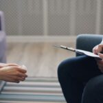 A counselor holds a clipboard and talks to a client.