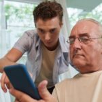 A young man helping an older man use a phone.