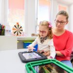 An adult woman holding up a child as they use an augmentative and alternative communication device.