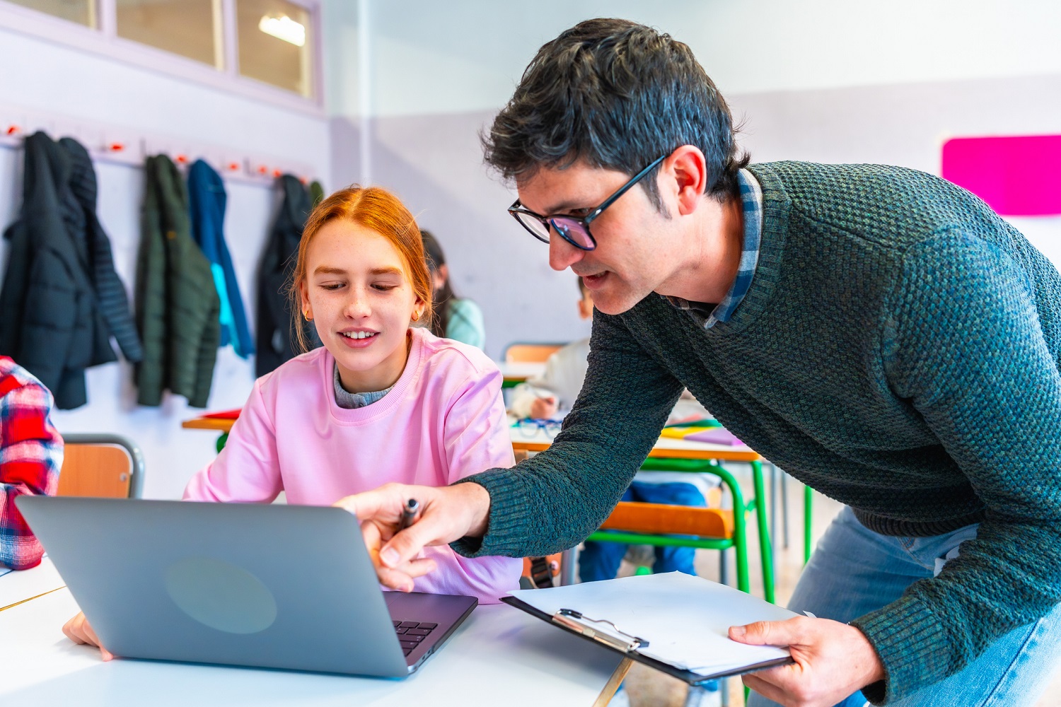 Teacher leaning over to assist a student working on a laptop in a bright classroom setting.