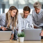 Three people in a workplace look over a laptop.