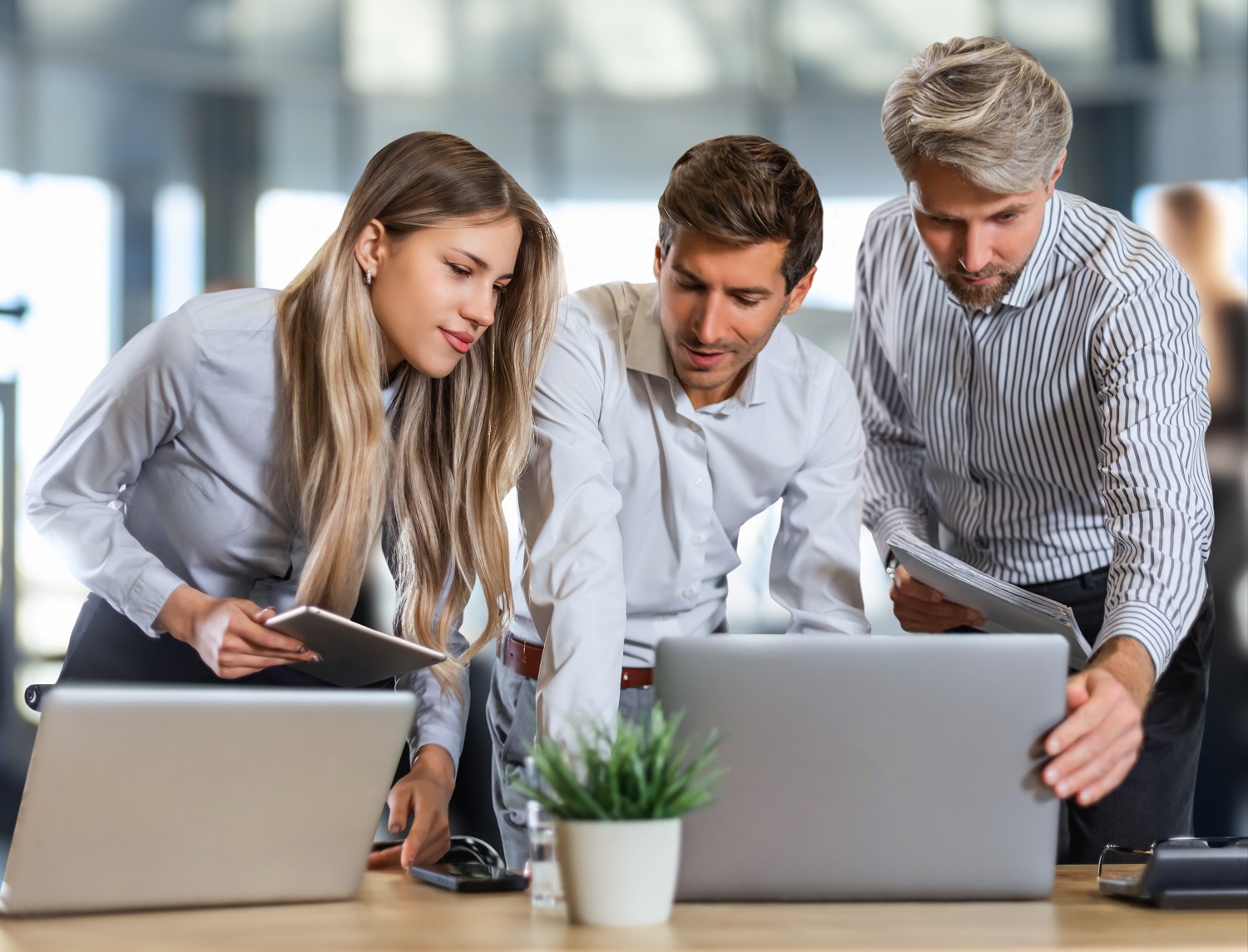 Three colleagues collaborating over laptops and tablets in a modern office setting with natural light.