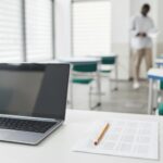 A desk with a laptop and a test in a classroom, with a teacher in the background a few rows back.
