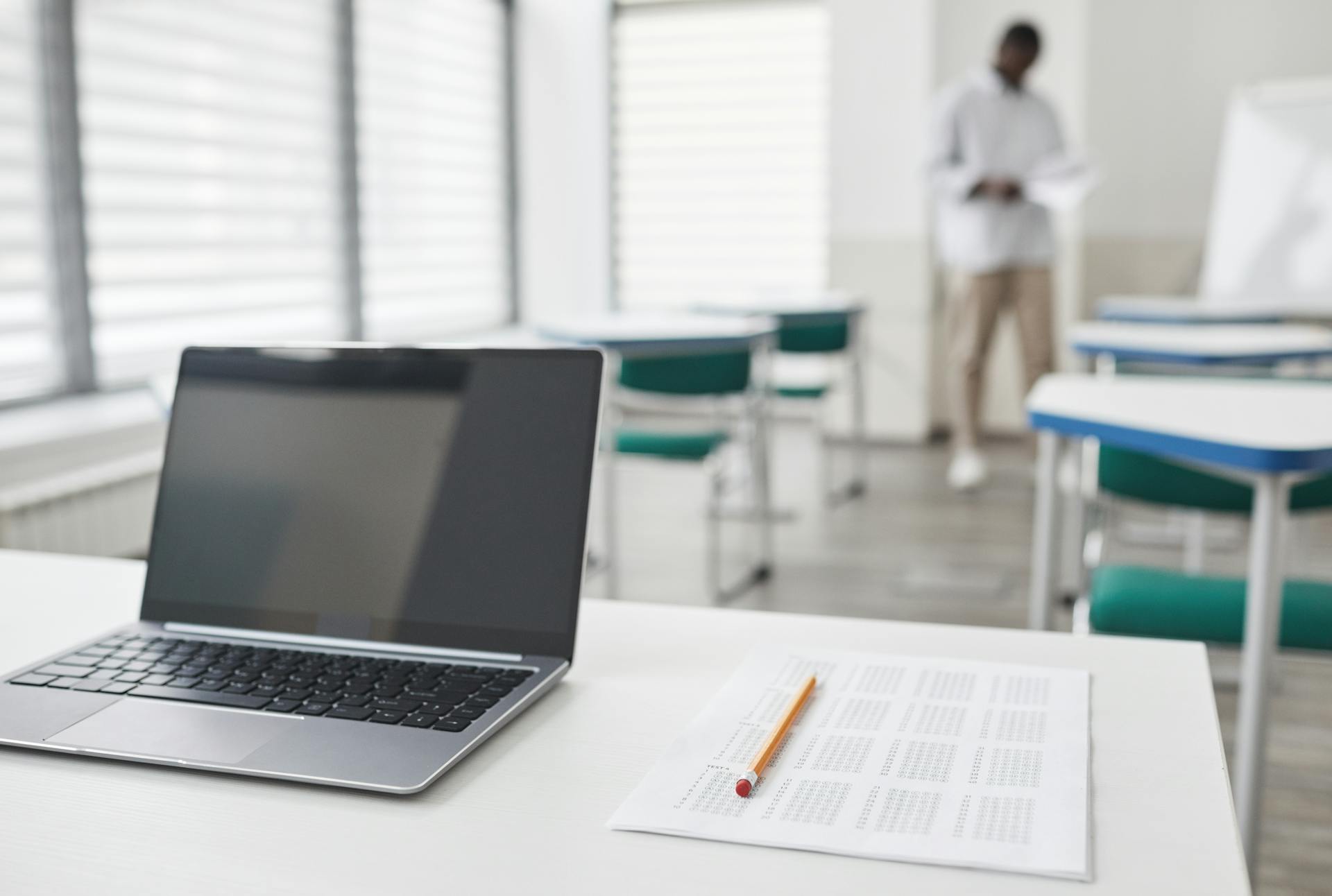 A desk with a laptop and a test in a classroom, with a teacher in the background a few rows back.