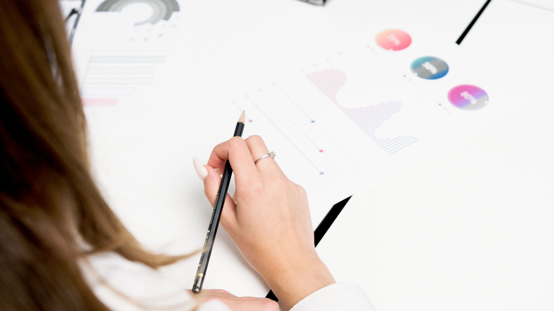 Person holding a pencil and analyzing printed charts and graphs on a white desk.