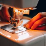 Close-up of hands guiding fabric through a sewing machine needle with warm lighting highlighting the stitching process.