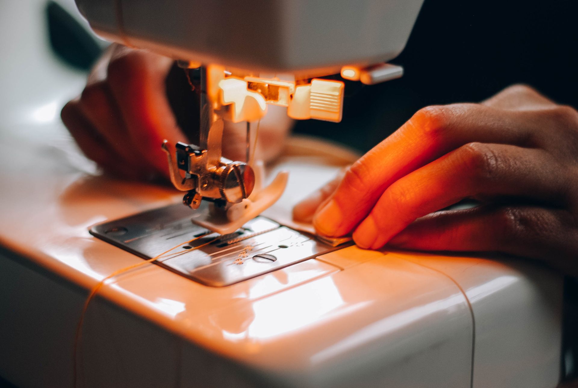 Close-up of hands guiding fabric through a sewing machine needle with warm lighting highlighting the stitching process.
