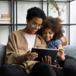 Smiling mother and daughter sitting on a couch together, enjoying a tablet screen in a cozy living room.